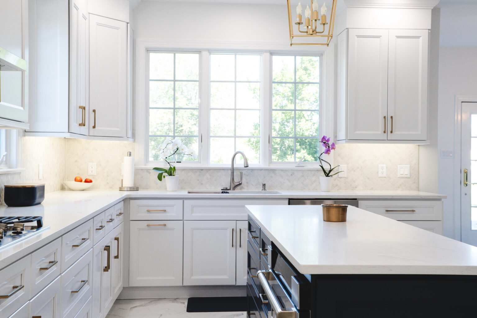 White kitchen with a dark island, broad window, gold-tone pulls, sink, orchids, and pendant light.
