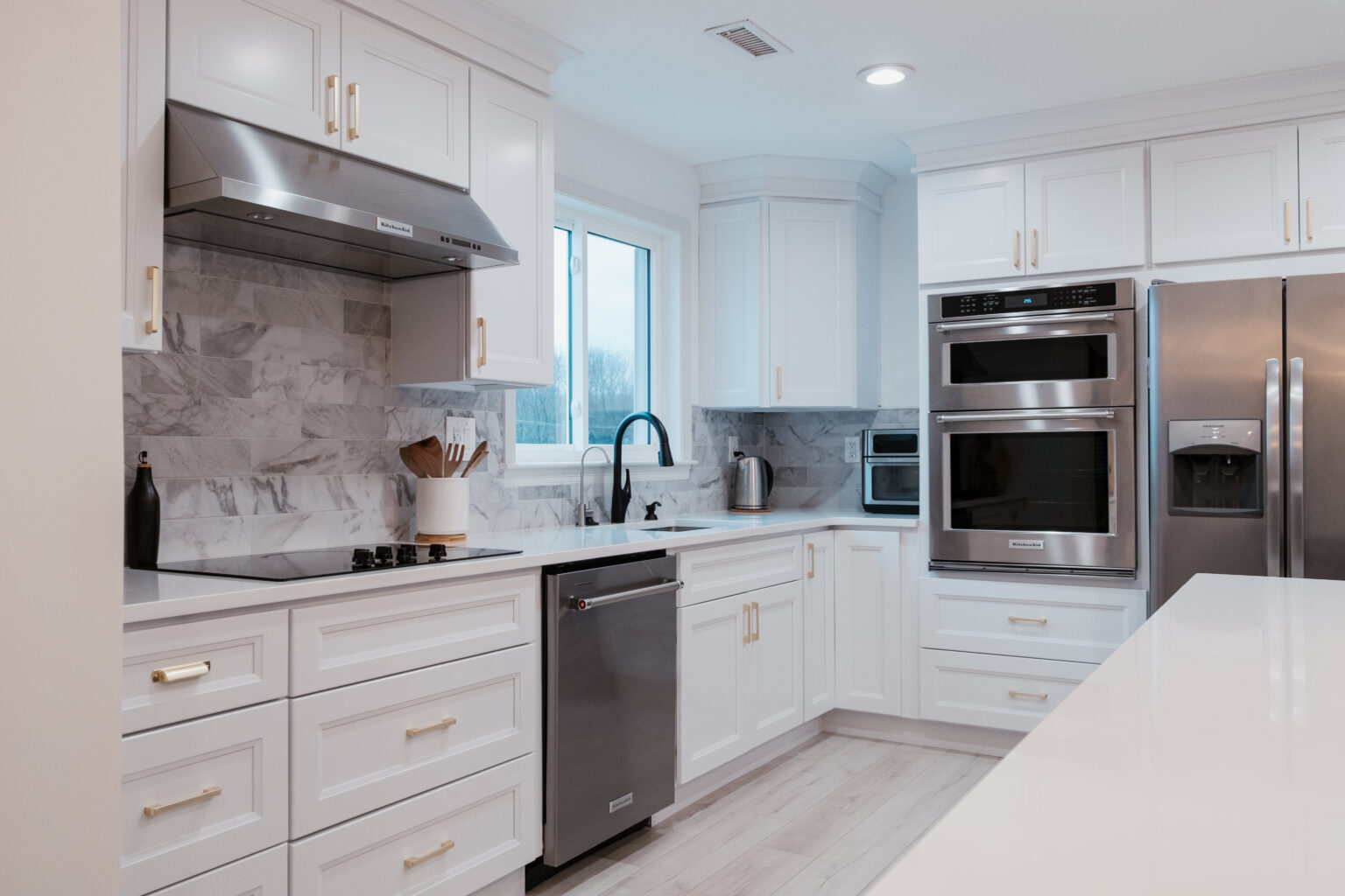 White kitchen with gold-tone pulls, veined backsplash, black faucet, and silver appliances.